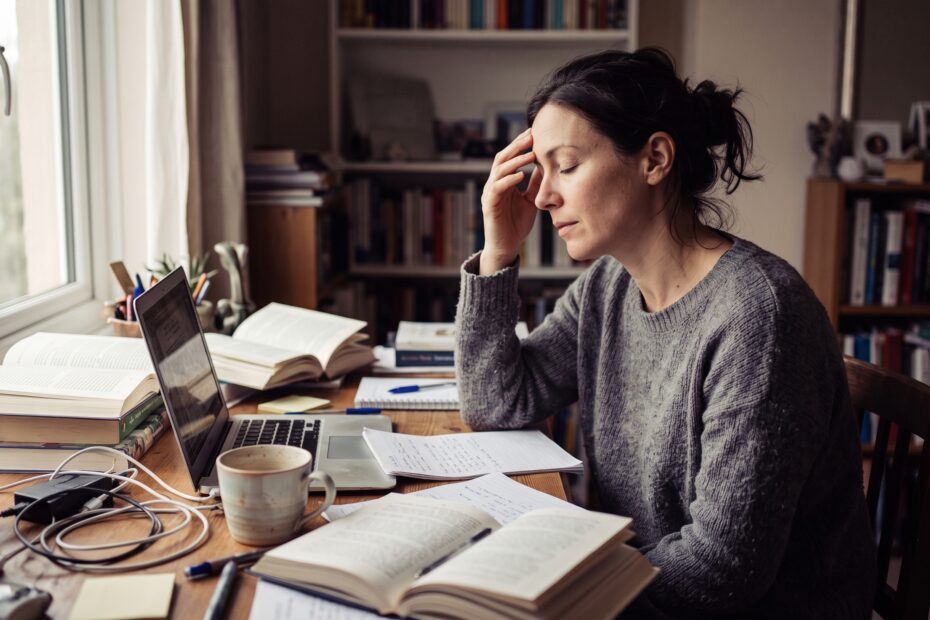 A person pausing at a cluttered desk in soft daylight, tired but composed, taking a breath.