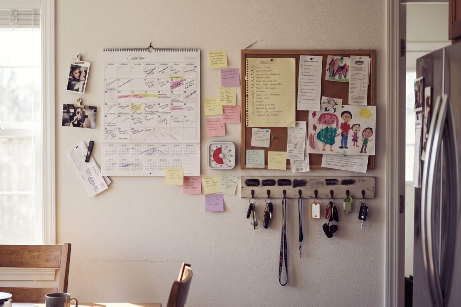 A kitchen wall command center with a paper calendar, sticky notes, a visual timer, and keys on a labeled hook in morning light.